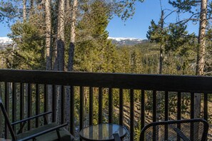 A wooden deck with a glass table and chairs, overlooking a forested area with a mountain in the distance.