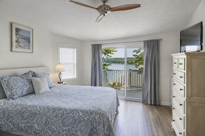 A bedroom with a bed, a ceiling fan, a dresser, a window with blinds, and a view of a lake.