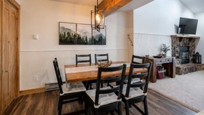 A dining area with a wooden table and chairs, a painting above the table, and a stone fireplace with a TV on a shelf. - Mountain Views from WP Top Floor Getaway (Winter Park)
