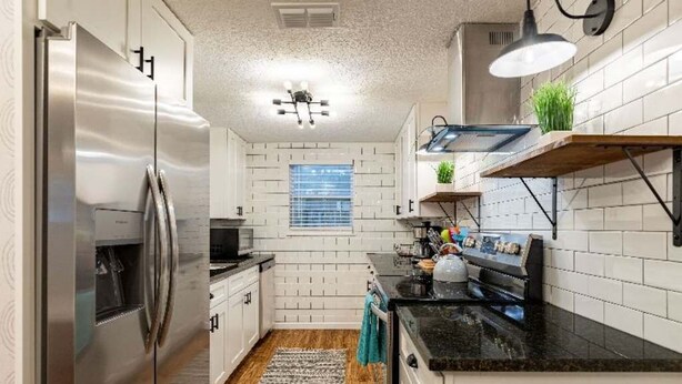 Una cocina moderna con armarios blancos, un refrigerador de acero inoxidable, una encimera negra y una lámpara colgante.