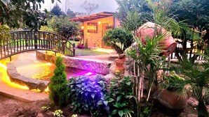 A garden with a stone bridge, a pond with illuminated edges, and a brick house in the background.