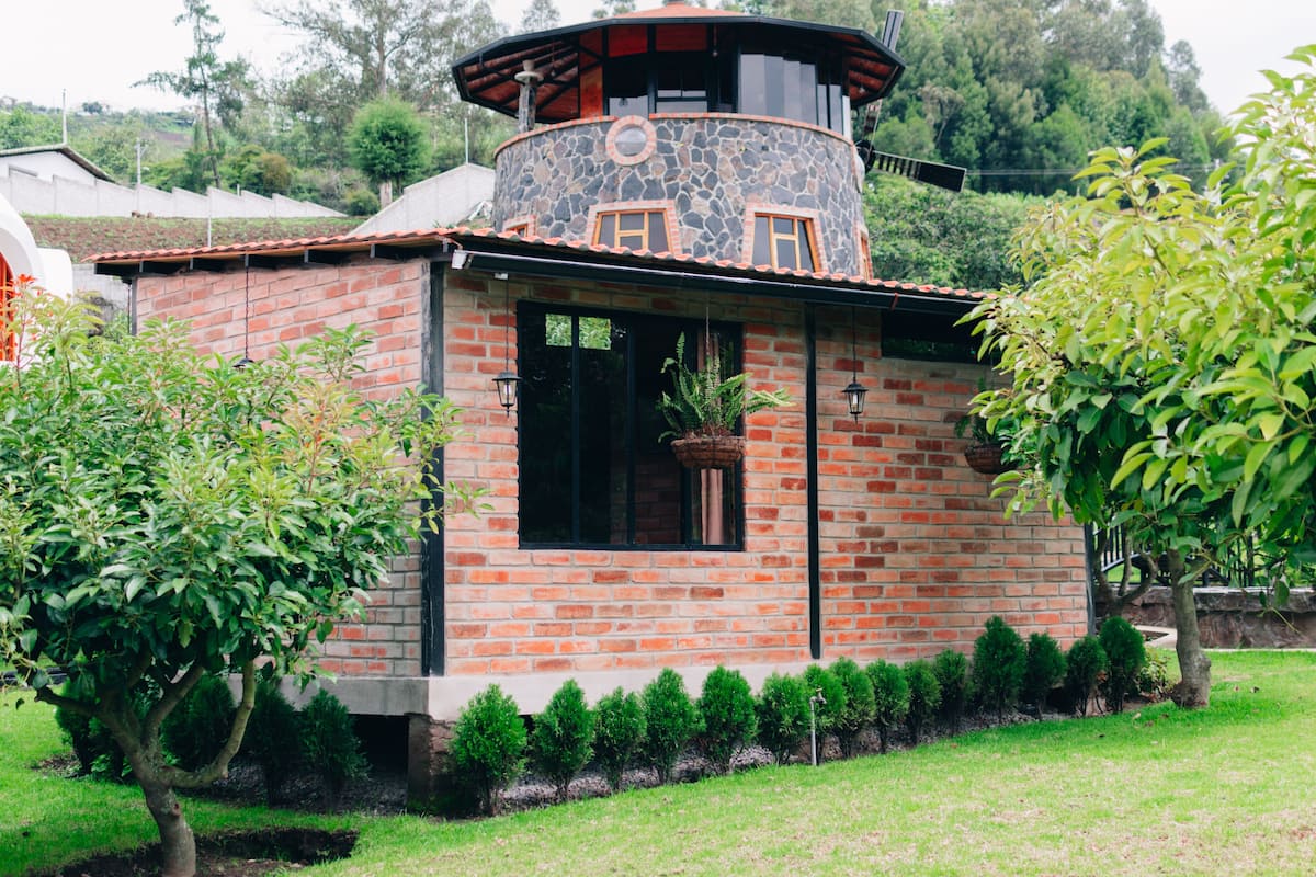 Edifici de maó amb una torre circular, una finestra amb planter penjant i un petit jardí amb arbustos podats.