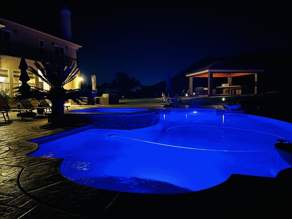 Una piscina con iluminación azul durante la noche, rodeada de mobiliario exterior y un edificio con porche cubierto.