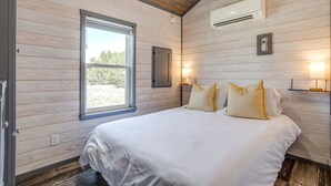 A bedroom with a bed, window, wall-mounted air conditioning unit, and bedside lamps.
