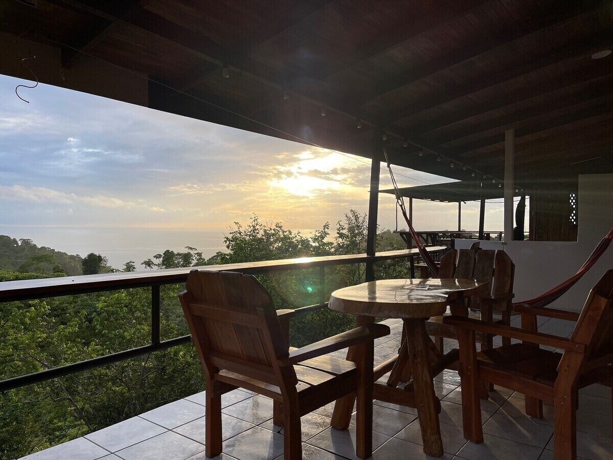An outdoor patio with a round table and wooden chairs, overlooking a lush landscape and the ocean.