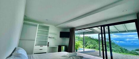 A modern bedroom with a large bed, a wardrobe, a television, and a balcony with a scenic view.