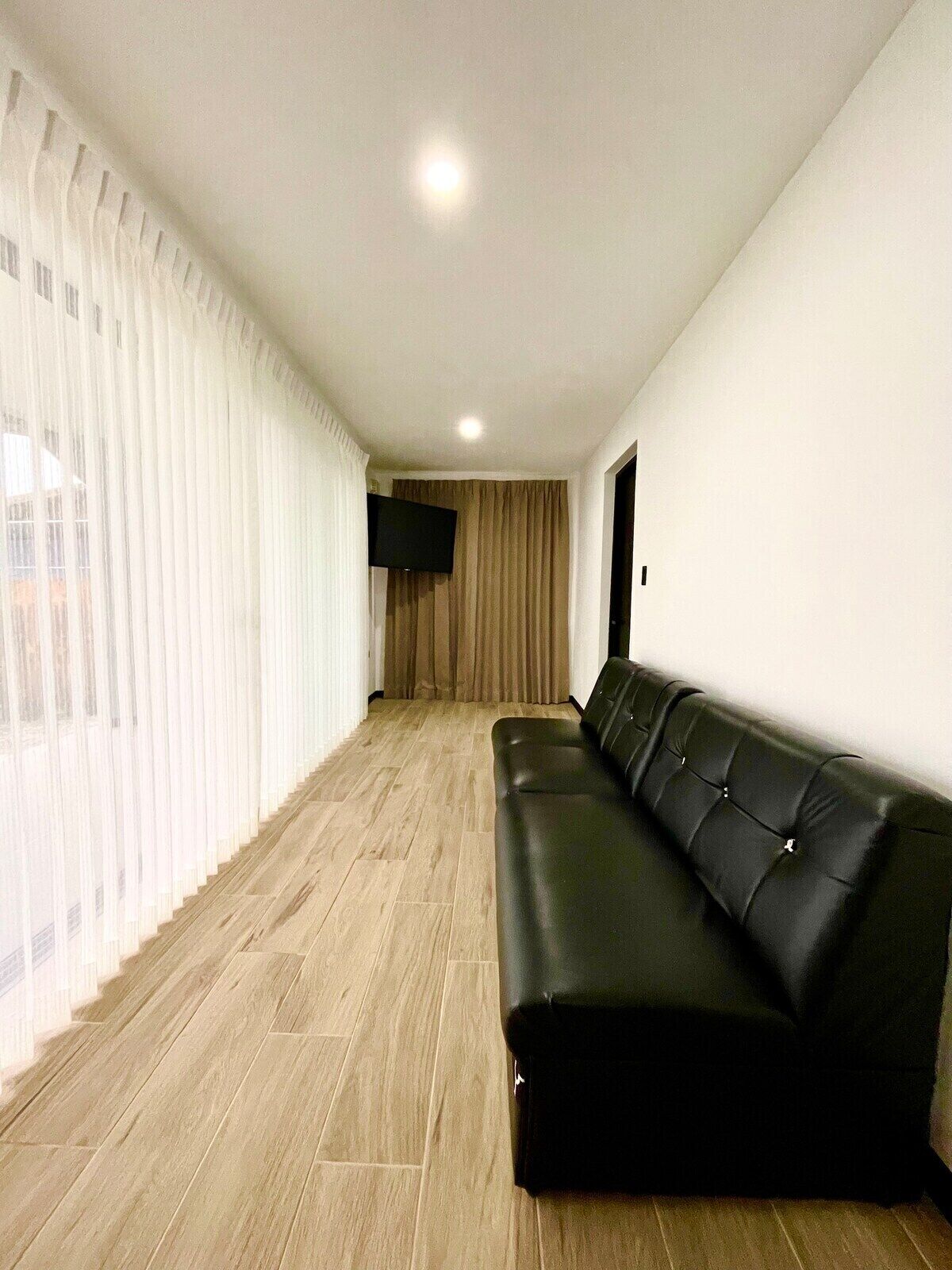 Uma sala de estar moderna com um sofá de couro preto, uma televisão de tela plana e uma janela grande com cortinas leves.