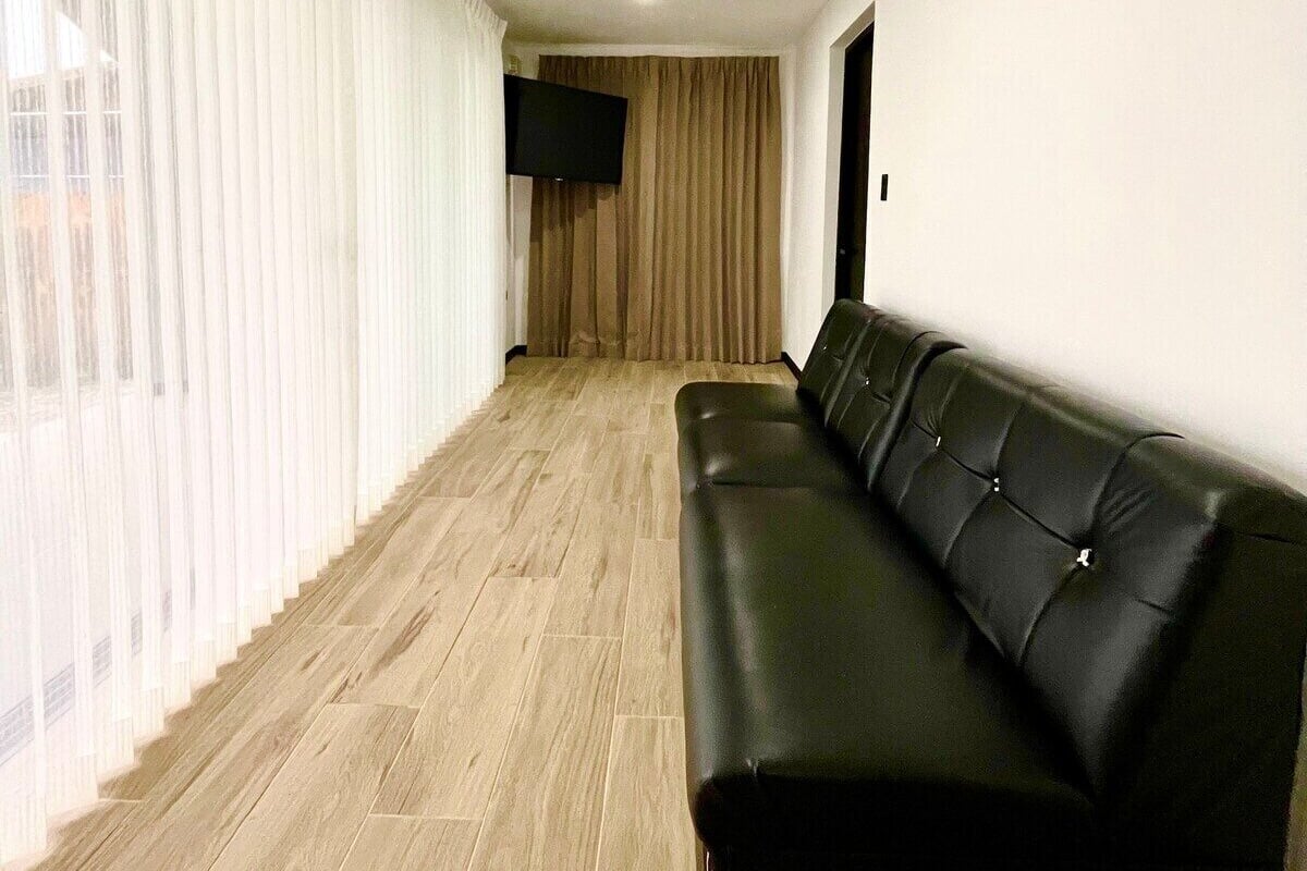 Uma sala de estar moderna com um sofá de couro preto, uma televisão de tela plana e uma janela grande com cortinas leves.