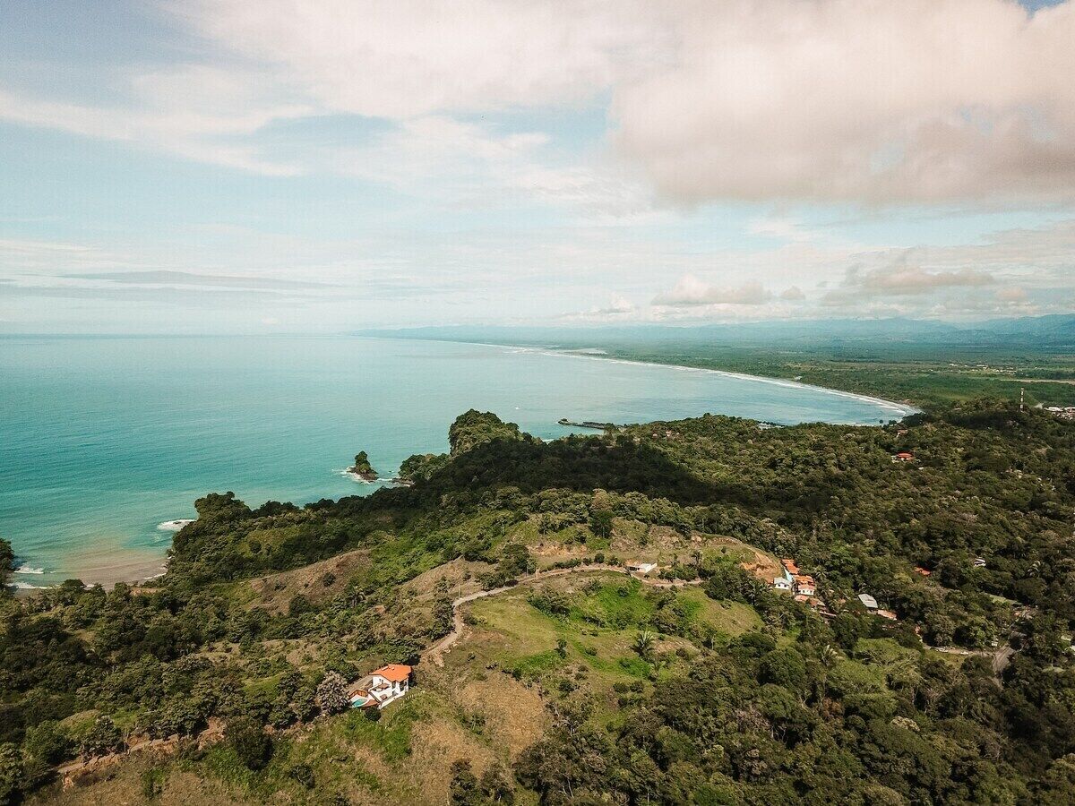 Vista aérea de una zona costera con un pequeño grupo de edificios rodeados de vegetación densa y una vista despejada de la costa.