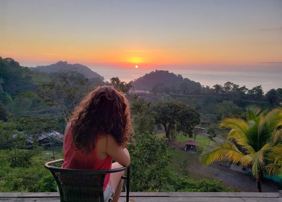A person seated on a wicker chair overlooking a scenic landscape with a sunset, hills, and a body of water.