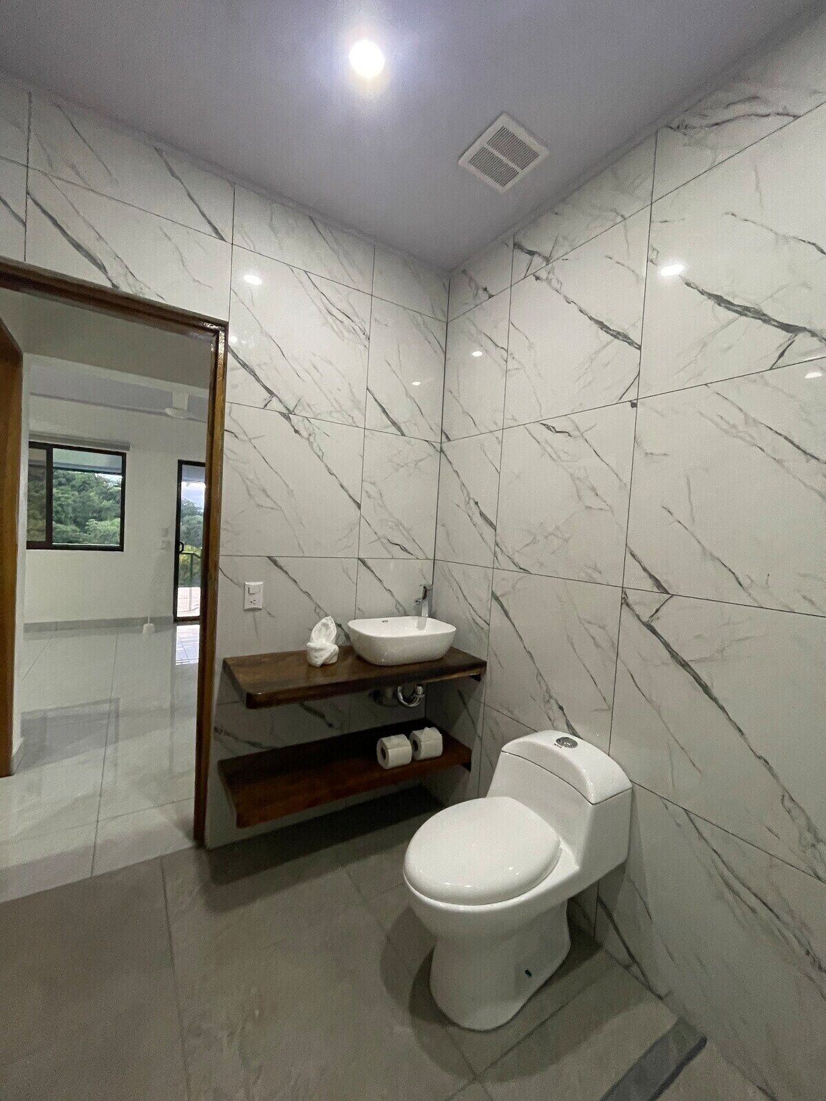A modern bathroom with a marble wall, a white toilet, a wooden vanity with a sink, and a mirror.
