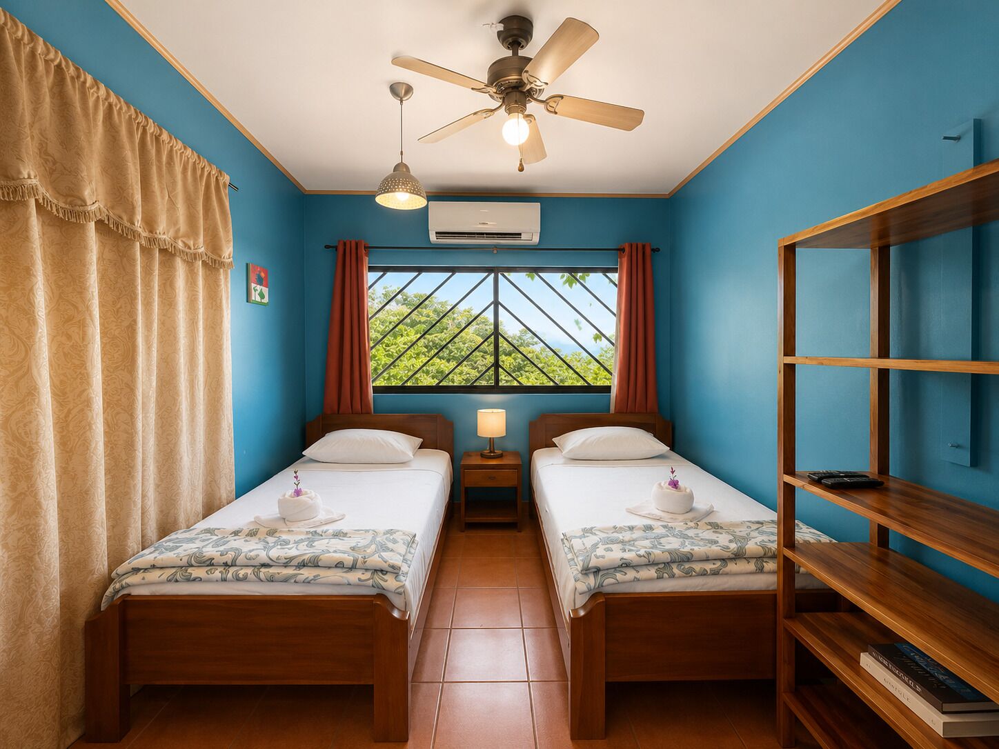 Una habitación con dos camas, ventilador de techo, unidad de aire acondicionado, ventana con vistas y una estantería.