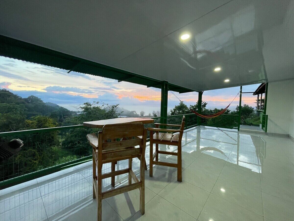 Un balcón con dos sillas de madera y una mesa, con vistas a un paisaje pintoresco con montañas y un atardecer.