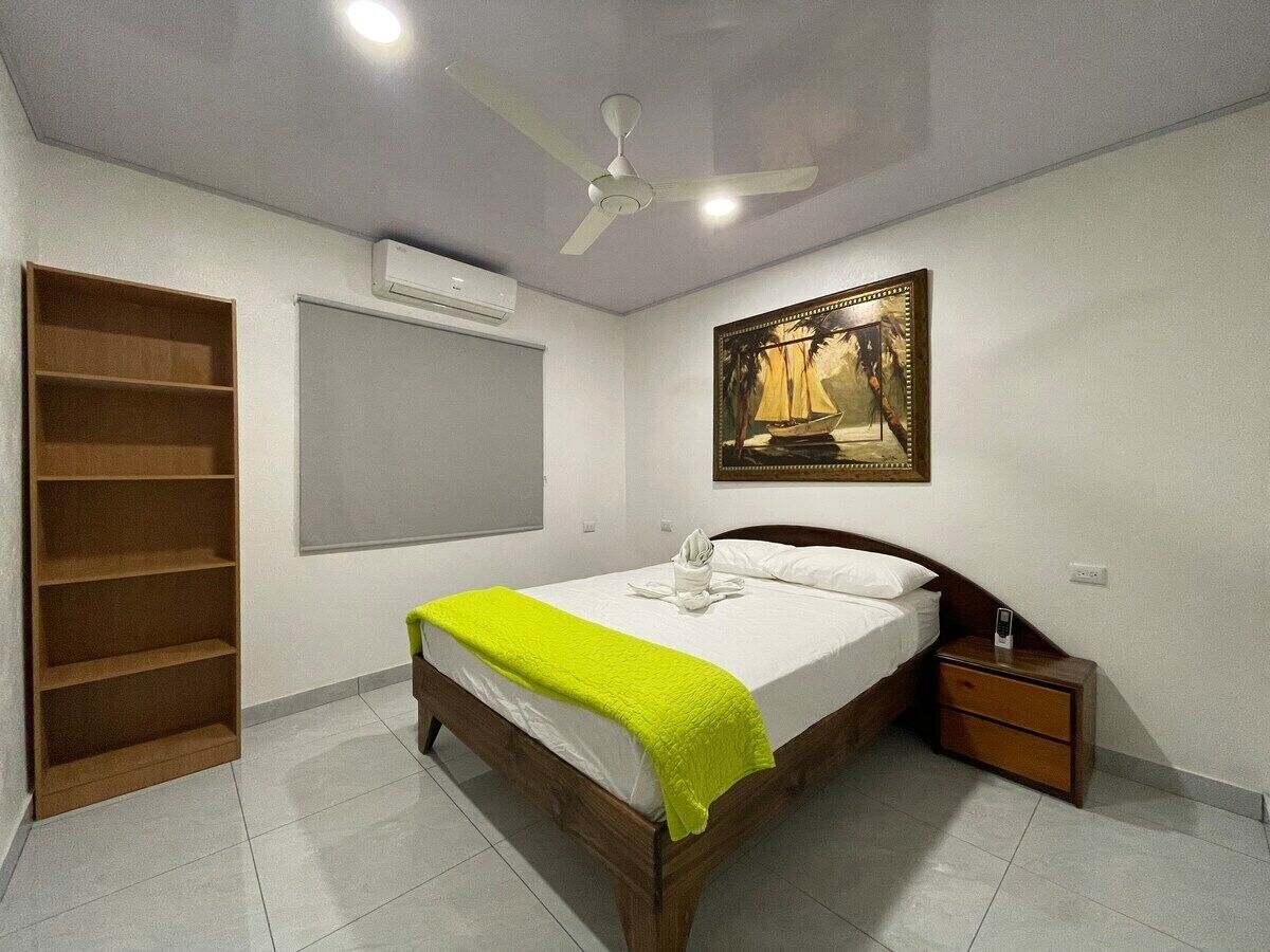 A bedroom with a bed, a nightstand, a wooden shelf, an air conditioning unit, and a ceiling fan.