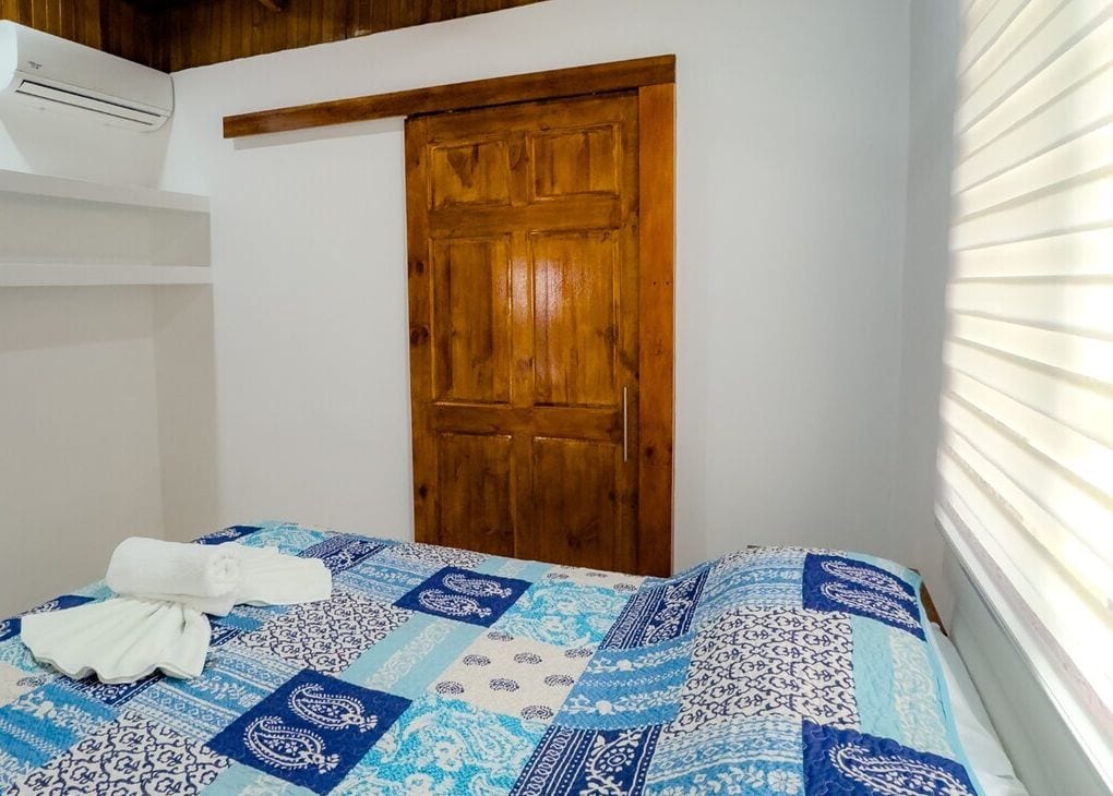 Quarto com porta de madeira, cama com colcha estampada em azul e branco, e um aparelho de ar condicionado na parede.