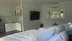 Une chambre dotée d’un grand lit, d’un téléviseur fixé au mur, d’une fenêtre donnant sur un espace verdoyant et d’un coin cuisine.