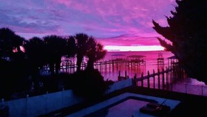 Une vue sur le coucher de soleil depuis un balcon donnant sur une étendue d’eau, avec une jetée en bois et des palmiers.