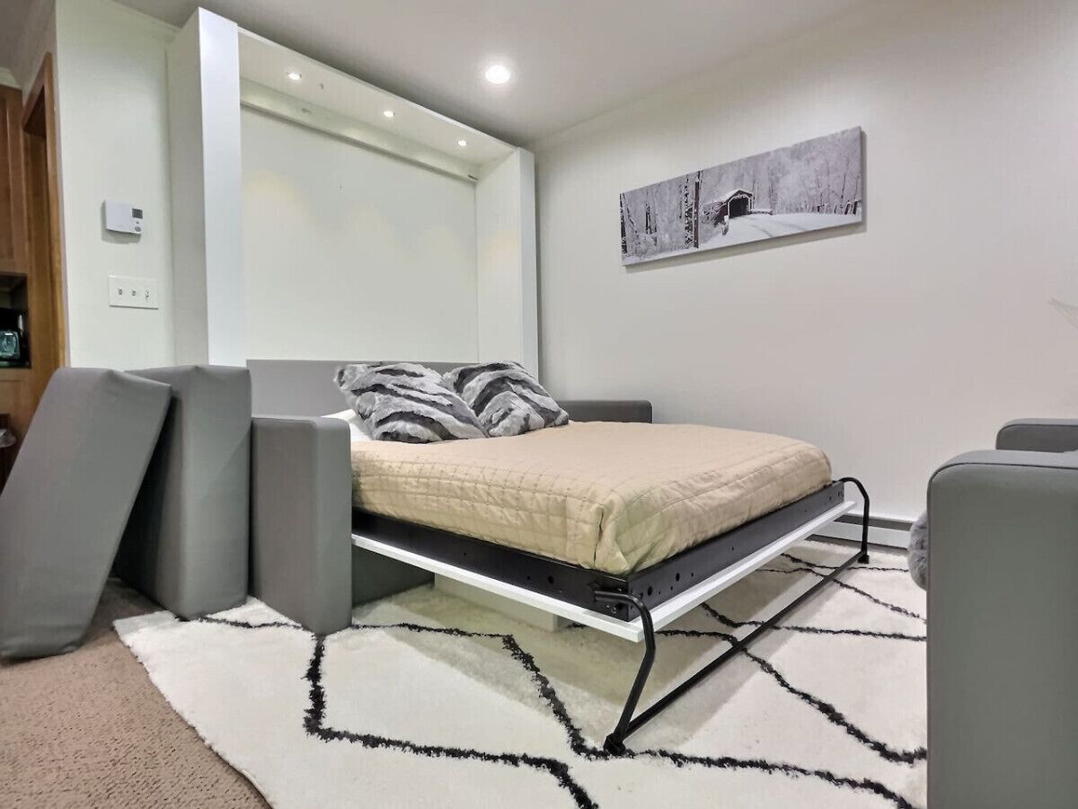 A compact bed with a mattress on a metal frame, a gray sofa, a white rug with a black pattern, and a wall-mounted artwork depicting a snowy scene.