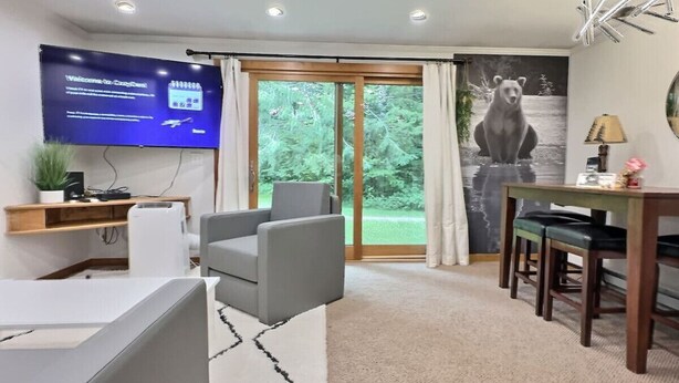 A modern living room with a flat-screen TV, grey sofas, a dining table with chairs, and a large window with a view of greenery.