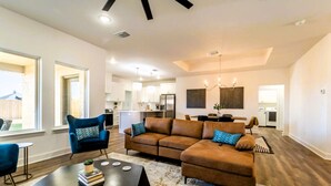 A spacious living room with a brown sectional sofa, a blue armchair, and a coffee table. There is a kitchen in the background with white cabinets and a dining area. - Chic New Build - 5 miles to Tech/Covenant/UHC - 2 miles to shopping! (Lubbock)