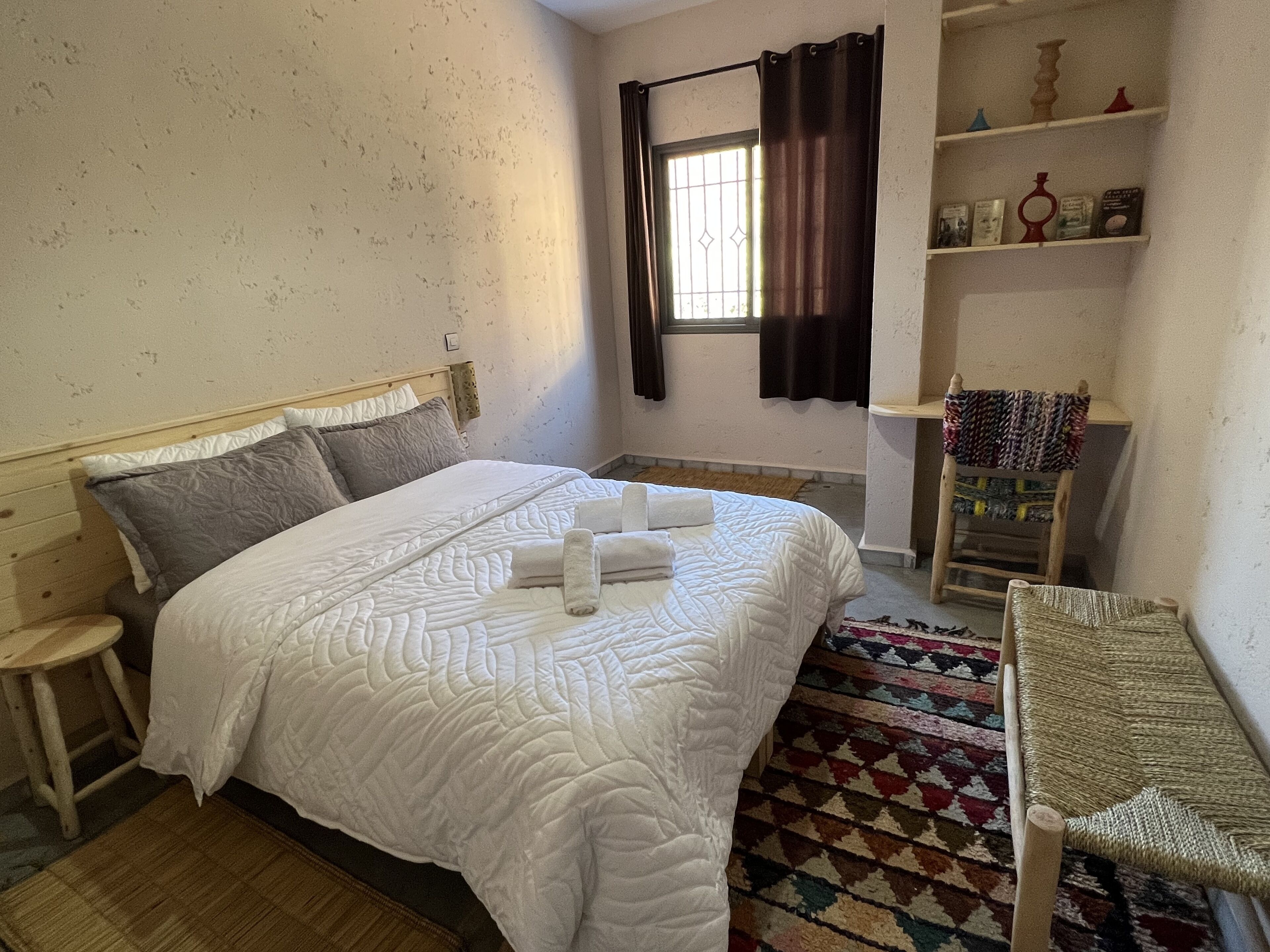 A bedroom with a bed, a small stool, a chair, a shelf with decorative items, and a window with curtains.