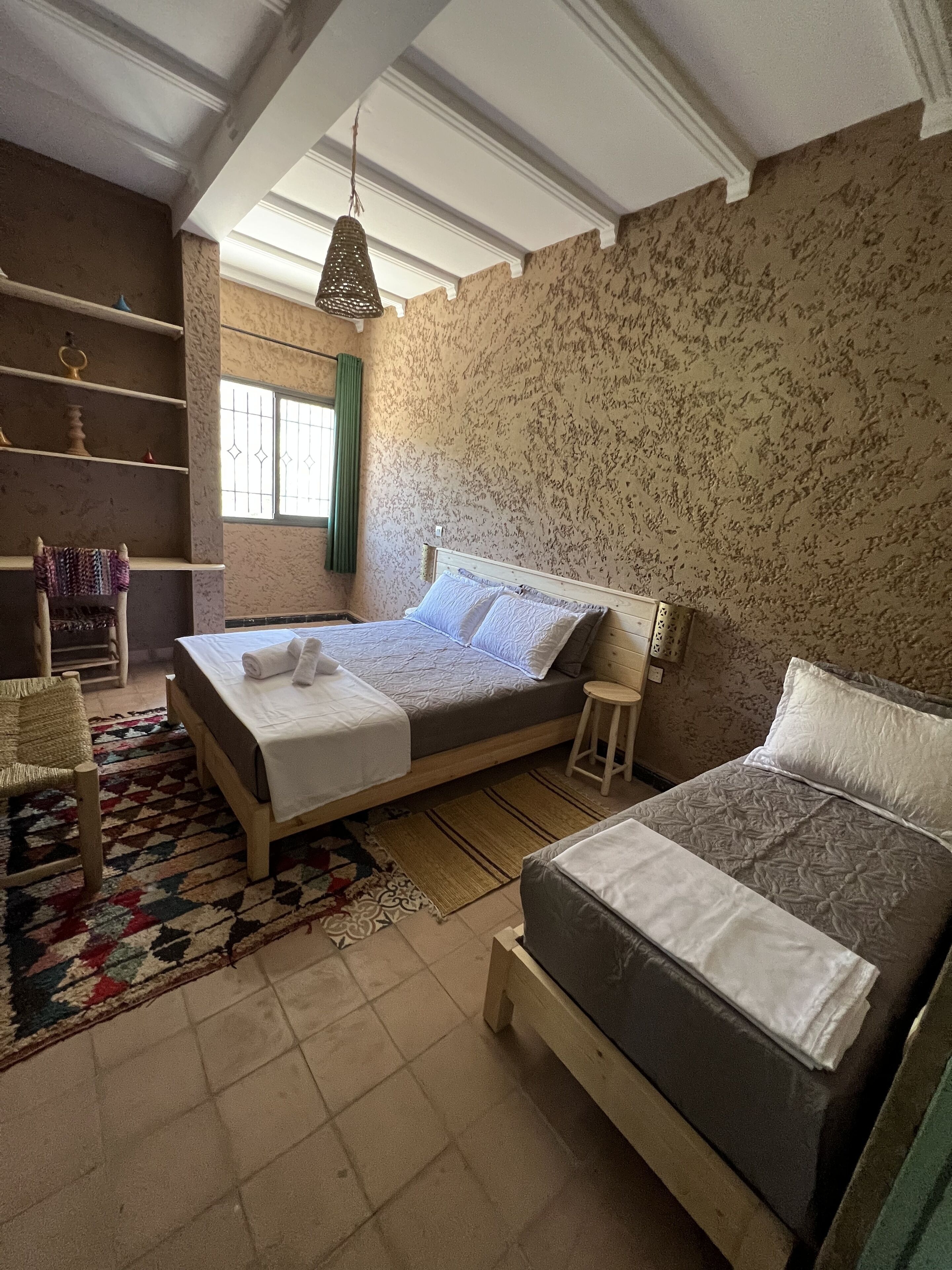 Ein Schlafzimmer mit zwei Betten, einem Holzstuhl, einem kleinen Hocker, einem gemusterten Teppich und einem Fenster mit Vorhängen.