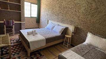 Ein Schlafzimmer mit zwei Betten, einem Holzstuhl, einem kleinen Hocker, einem gemusterten Teppich und einem Fenster mit Vorhängen.