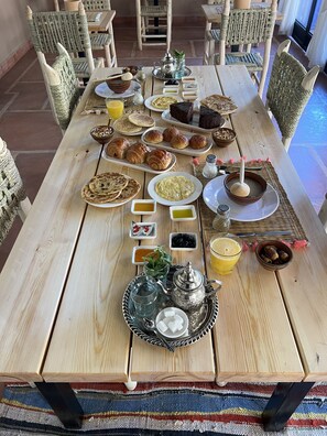 A breakfast buffet with various dishes, including pastries, bread, and beverages, set on a wooden table in a cozy dining area with wicker chairs. - Tazart Lodge (Sti Fadma)