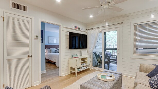Um quarto bem iluminado, com ventilador de teto, televisão montada na parede, porta de vidro deslizante que dá para uma área externa e um espaço de estar acolhedor com sofá e apoio para os pés.