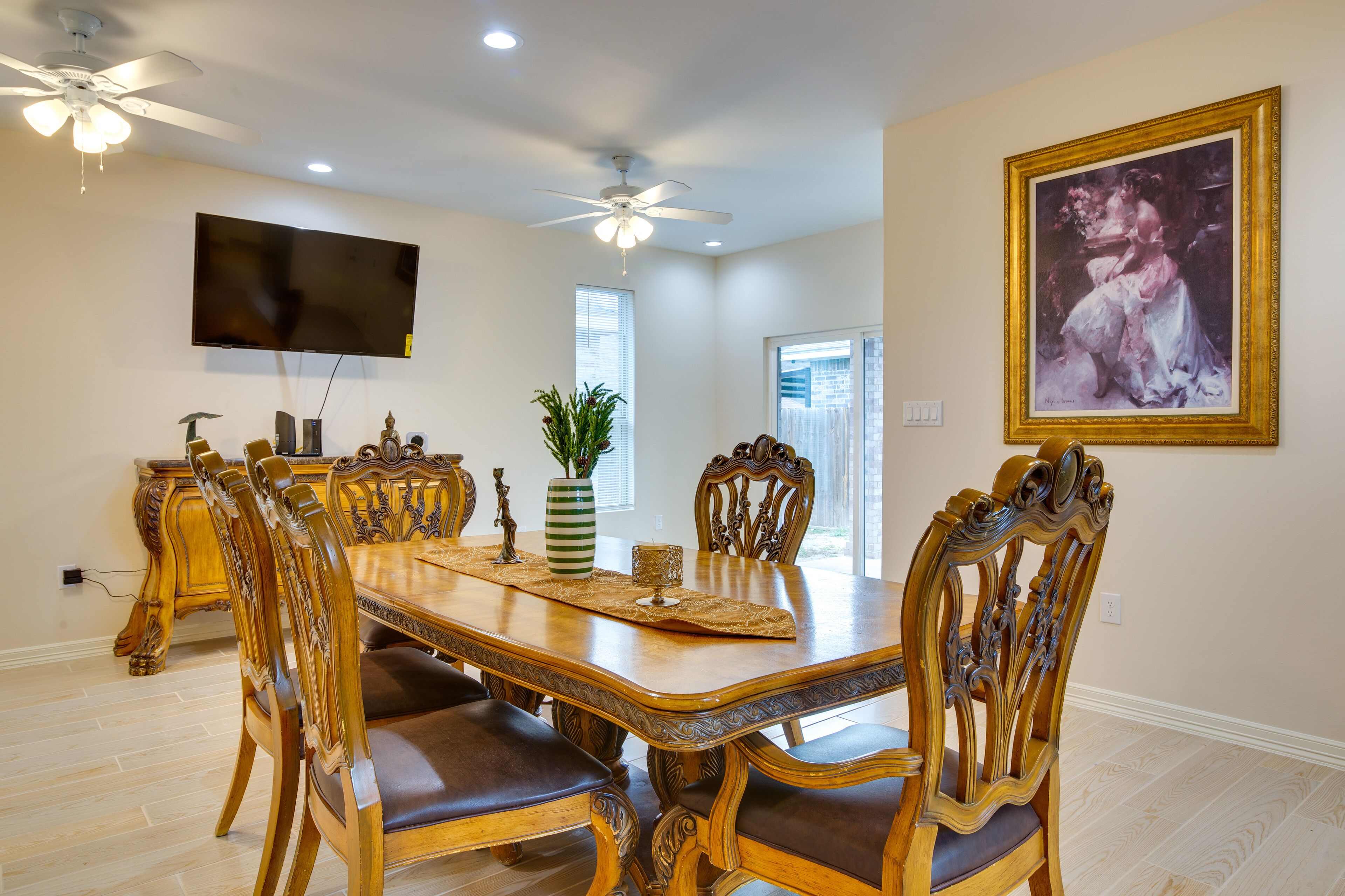 A dining room with a large wooden table, ornate chairs, a ceiling fan, a television, and a framed painting on the wall.