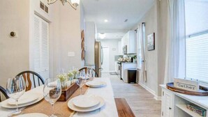 Un coin repas avec une longue table dressée pour quatre personnes, une cuisine en arrière-plan et un panneau indiquant «Rassemblement» placé sur une étagère.