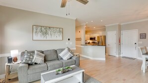 Una sala de estar moderna con un sofá gris, una mesa de centro blanca y un ventilador de techo. Se aprecia una cocina al fondo con armarios blancos y electrodomésticos de acero inoxidable.