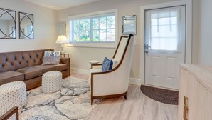 A living room with a brown leather sofa, two patterned ottomans, a light-colored armchair, a small side table with a lamp, a large window with blinds, and a white door. - 2 Blocks to Riviera Beach: Upscale Condo w/ Patio (West Palm Beach)
