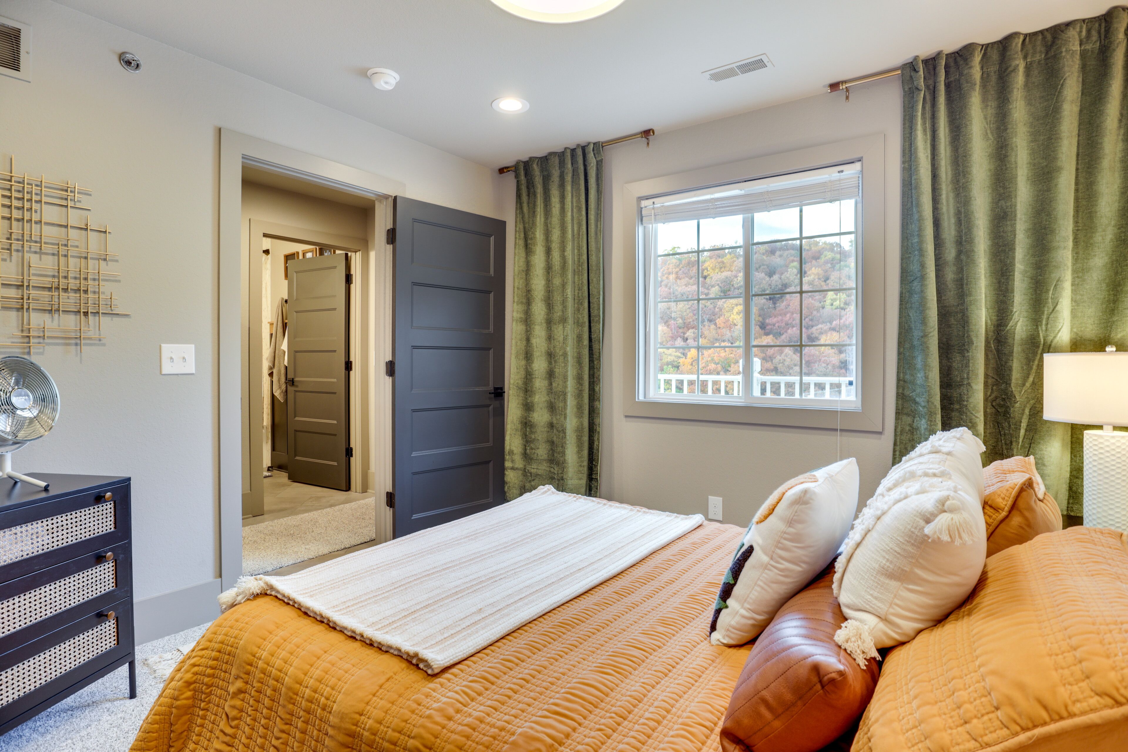 A bedroom with a bed, a bedside table, a lamp, a window with curtains, and a door leading to another room.