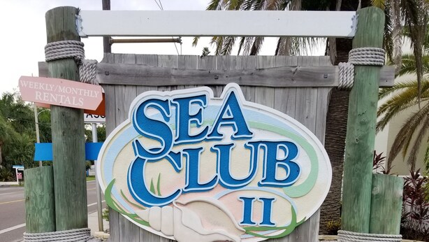 Uma placa de madeira com o nome Sea Club II e o número '5955' abaixo, rodeada por um canteiro de flores.