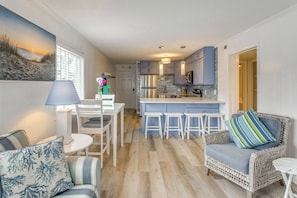 A modern kitchen with a central island, bar stools, and a wicker sofa with a striped pillow. - Steps to Ocean: High-End North Myrtle Beach Condo (North Myrtle Beach)