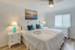 Une chambre à coucher avec un lit, deux tables de chevet avec des lampes, un ventilateur de plafond, une fenêtre avec des stores et un tableau au mur.