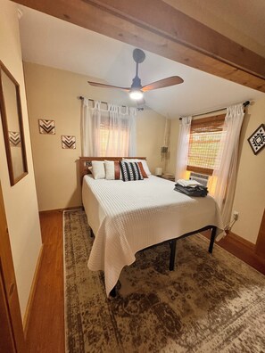 A bedroom with a bed, a ceiling fan, a window with curtains, a wall-mounted air conditioning unit, and a patterned rug.