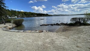 Ein sandiger Strand mit felsigem Ufer, ein ruhiger See und eine ferne Ansicht von Häusern und Bäumen.