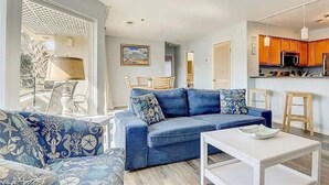 Una sala de estar moderna con un sofá azul, una mesa de centro blanca y una zona de cocina con armarios de madera y un microondas.
