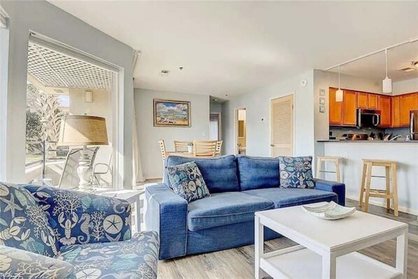 Una sala de estar moderna con un sofá azul, una mesa de centro blanca y una zona de cocina con armarios de madera y un microondas.