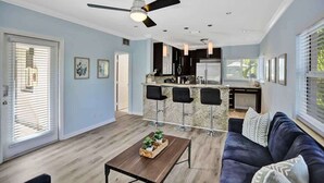 A modern kitchen with bar stools, a ceiling fan, and a dining area with a dark-colored sofa and a coffee table. - Modern 1BR on 2nd Floor Near Las Olas (Fort Lauderdale)