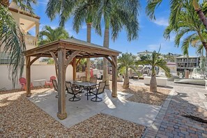 Een overdekt terras met een tafel en stoelen, omgeven door palmbomen en een heldere lucht.