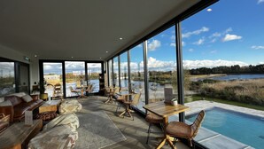 Un espace intérieur moderne doté d’une grande fenêtre donnant sur un lac et des montagnes. On y trouve des coins salon avec des tables et des chaises, et une piscine est visible à l’extérieur.