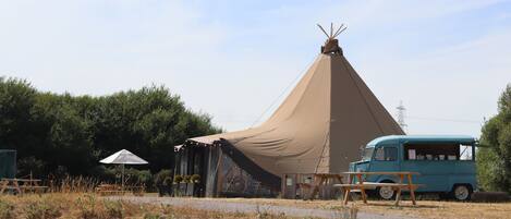Una tienda de campaña con techo cónico, una furgoneta turquesa antigua y mesas de picnic en un césped.