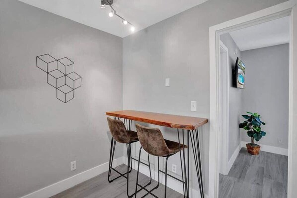 Un coin repas avec une table en bois et deux tabourets de bar, une œuvre d’art géométrique accrochée au mur et une plante en pot.