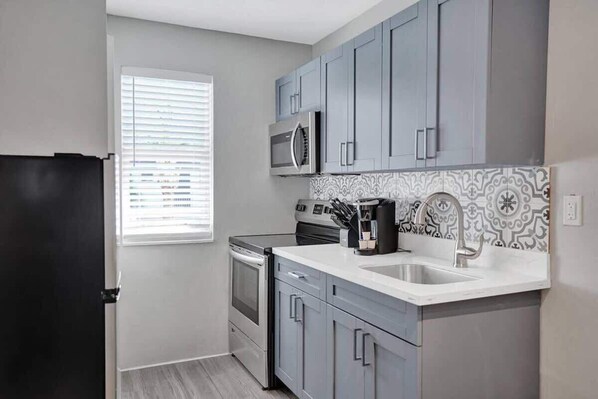 A compact kitchen with grey cabinets, a stainless steel refrigerator, a microwave, an oven, and a sink with a patterned backsplash.