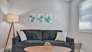 Een moderne woonkamer met een grijze bank, een ronde houten salontafel en een vloerlamp. Boven de bank hangen drie plantenprints op de muur.