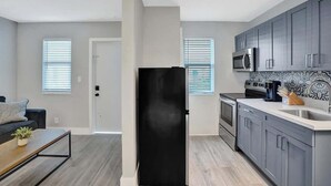 Una cuina moderna amb armaris grisos, un refrigerador negre i un microones. Hi ha una zona de menjador amb taula de fusta i un sofà.