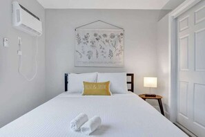 Una camera d'albergo con un letto, un condizionatore fissato al muro, un comodino con una lampada e un'illustrazione botanica incorniciata.